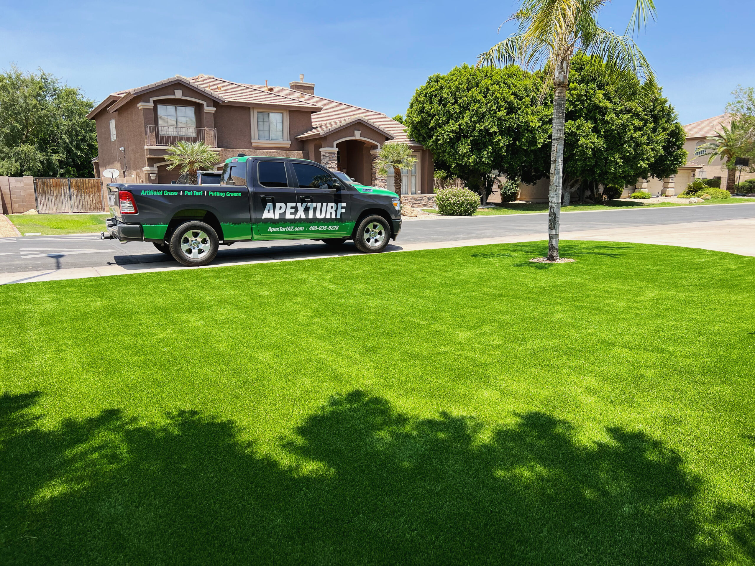 A vibrant green lawn is in the foreground with a pickup truck parked on the street. The truck, showcasing "APEXTURF" branding, hints at their expertise in artificial grass for Chandler. Suburban houses, trees, and a clear blue sky complete this idyllic scene.