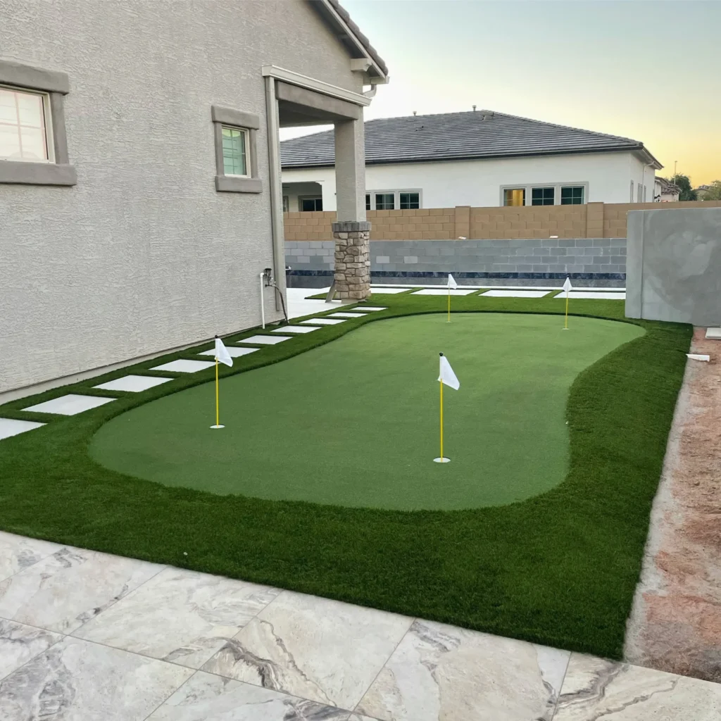 A small backyard putting green is bordered by fringe grass and stone pavers. Four white flags on yellow sticks are spaced at each hole on the golf green. A modern house and privacy wall are in the background under a clear sky.