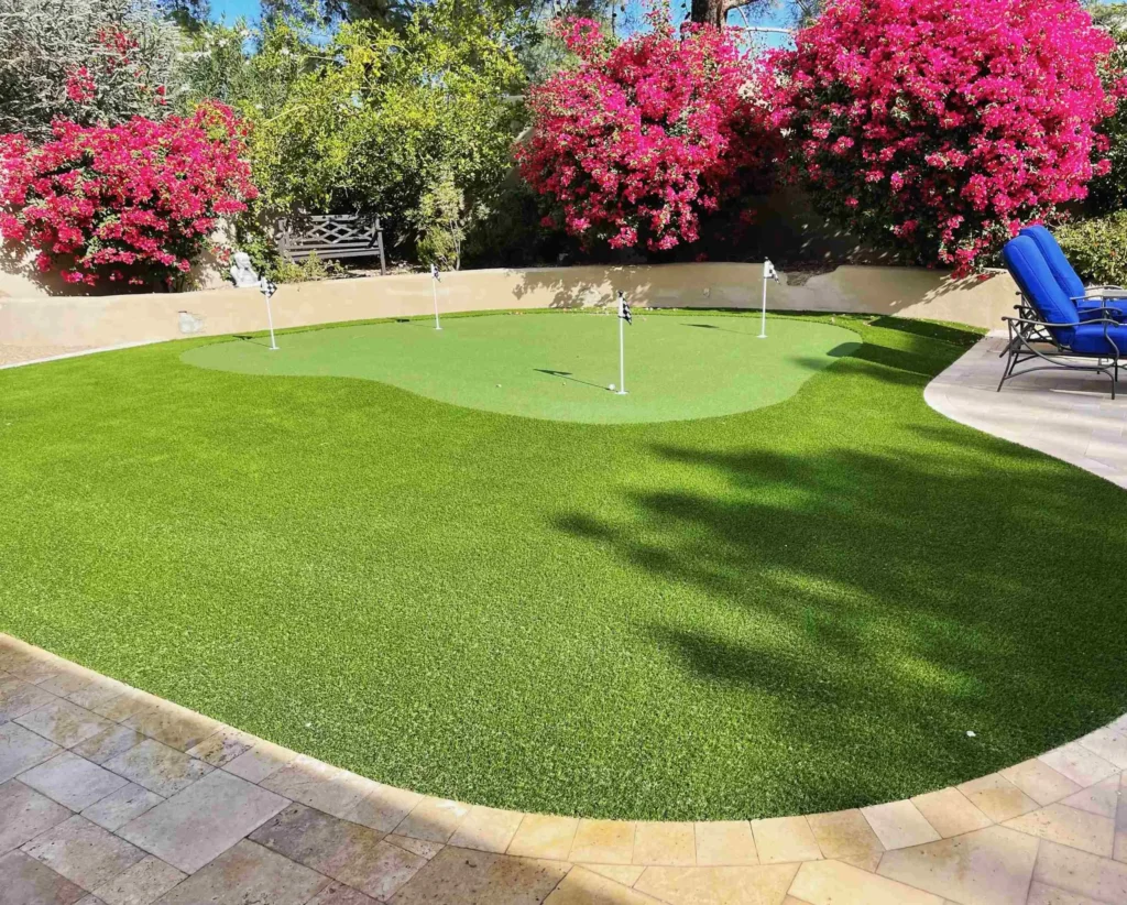 A backyard putting green of premium Apex Turf is surrounded by vibrant pink flowering bushes. The area is bordered by a patio with beige tiles, featuring two blue lounge chairs. The scene is sunny, casting delightful shadows on the lush green grass.