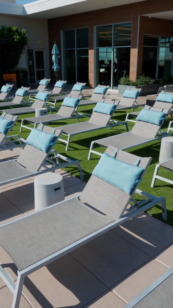 Rows of modern, gray lounge chairs with blue cushions grace a sunlit patio near a building adorned with large glass windows. The serene space, featuring small round side tables, is beautifully surrounded by lush artificial grass from Chandler's Apex Turf.