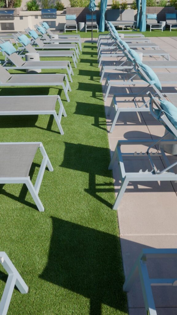 Sun loungers with gray frames and blue umbrellas are tastefully arranged on a rooftop, partly on artificial grass from Apex Turf and partly on concrete. Some loungers have blue cushions, casting elegant shadows—a serene escape high above Chandler's hustle.