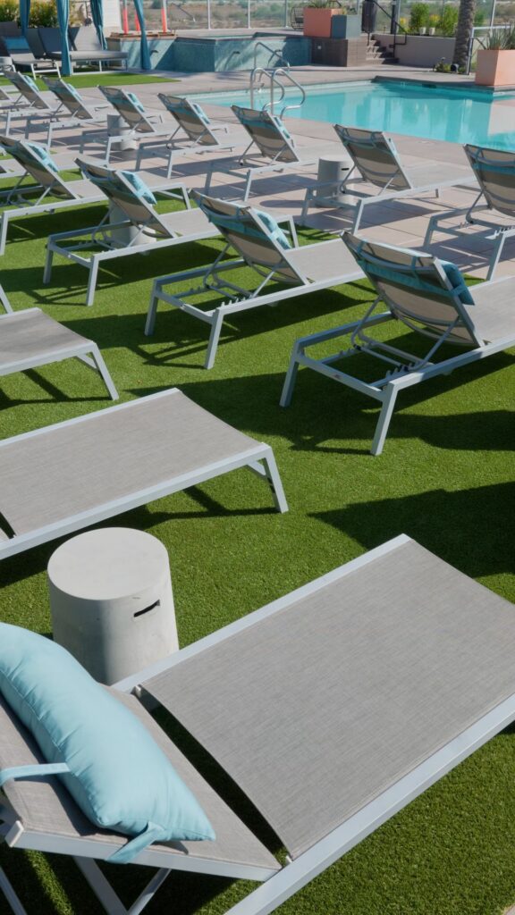 Several lounge chairs and small side tables are arranged on the lush artificial grass by Chandler's Apex Turf, near a swimming pool. Cushions on the chairs are light blue. The pool area is fenced, with palm trees and planters providing a serene backdrop.