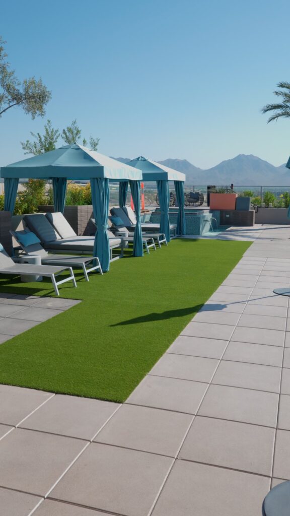 The lounge area features turquoise cabanas and sunbeds beside a vibrant green artificial lawn from Apex Turf. A paved walkway graces the foreground, with mountains majestically set against a clear blue sky overhead, capturing the essence of Artificial Grass Chandler.