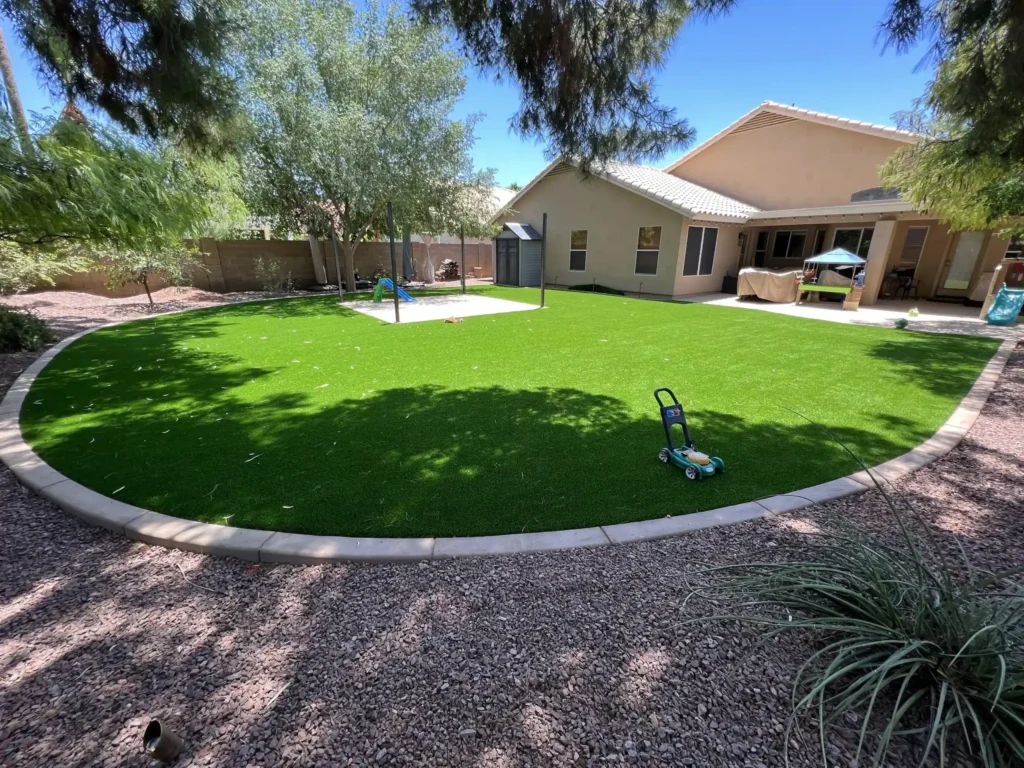 Backyard with green artificial grass, bordered by gravel and trees. A toy lawnmower sits on the grass. A small play structure and a tan house with a patio are in the background under a clear blue sky.