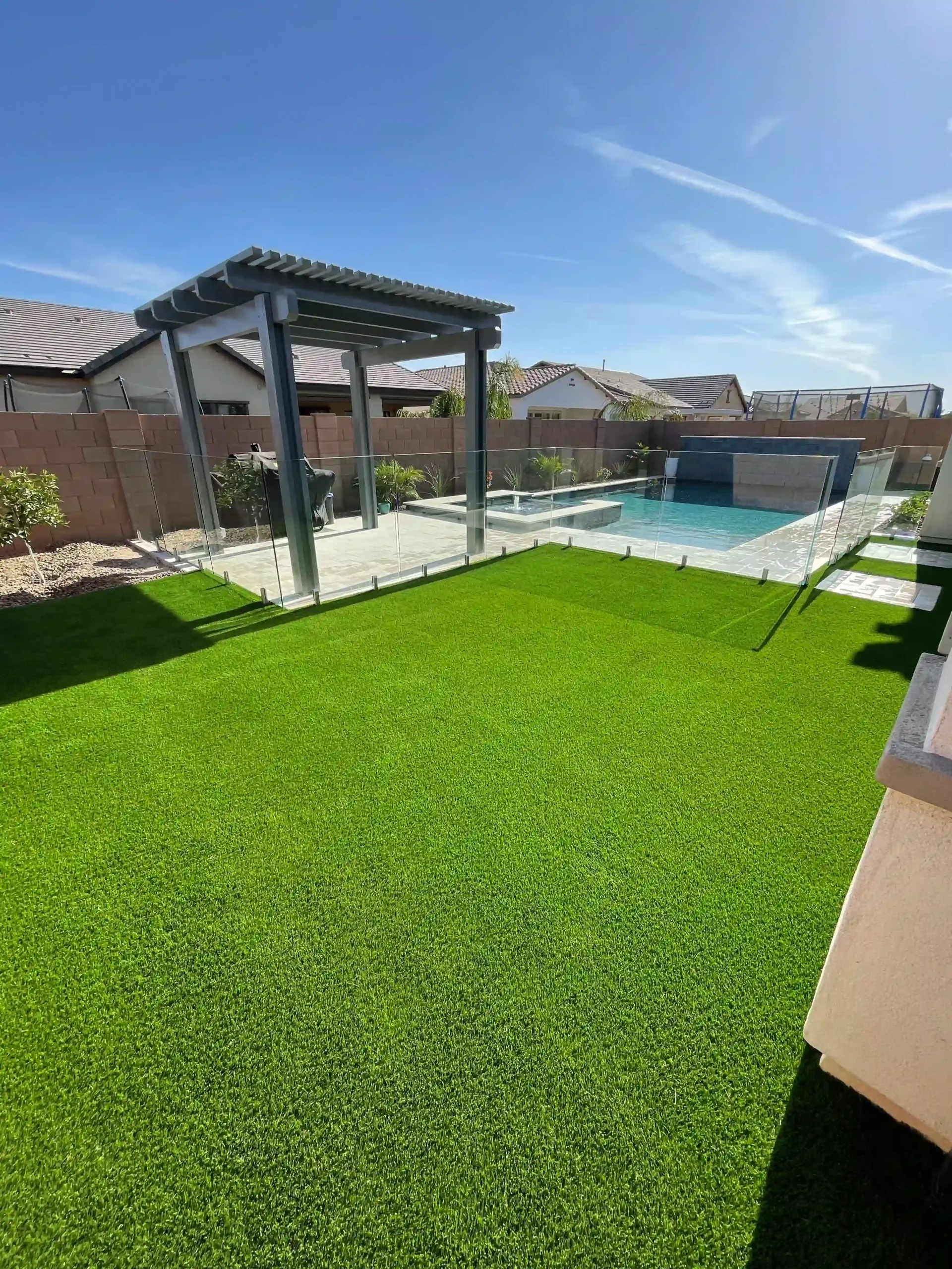 A backyard featuring Apex Turf's manicured artificial grass lawn, framed by a glass fence. A shimmering swimming pool and a pergola add charm in the background. The sky is clear and blue, adorned with a few wispy clouds.