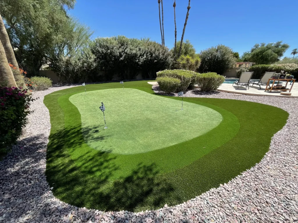 A lush, green Apex Turf putting green is surrounded by pebbled landscaping under a clear blue sky. Two small flags mark holes. Tall palm trees and various shrubs border the area, with patio furniture visible in the background.