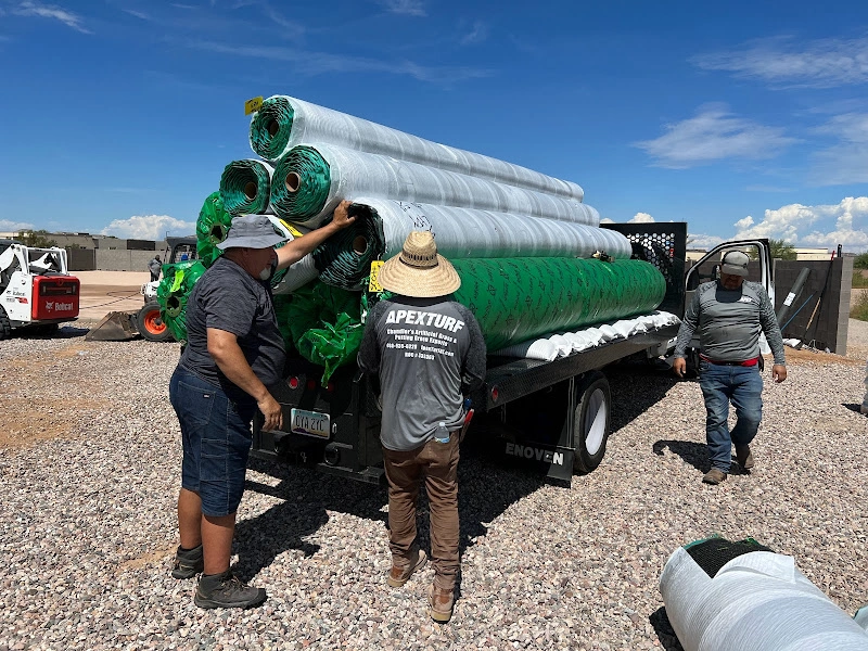 Three workers load large rolls of Apex Turf onto a black flatbed truck on a gravel surface. One person wears a straw hat and a shirt proudly displaying "Apex Turf." A blue sky and construction equipment fill the background.