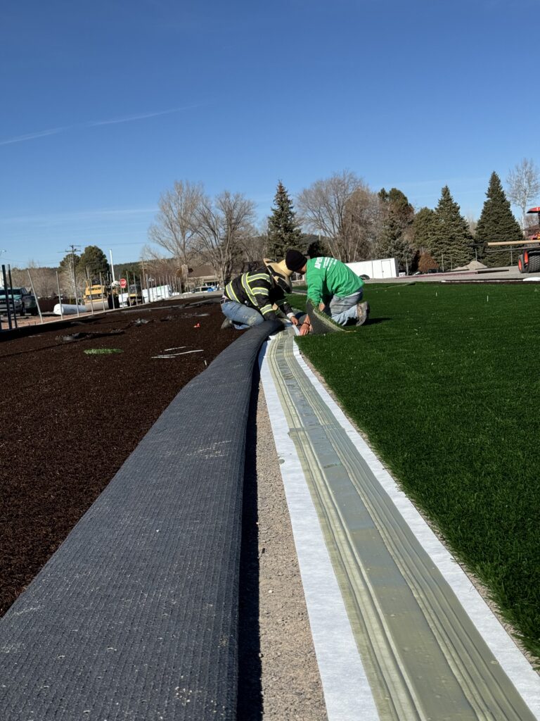 Two workers in safety gear install synthetic turf on a sunny day. They kneel on the ground, aligning and securing the green grass carpet on a sports field, with barren trees and clear blue sky in the background.