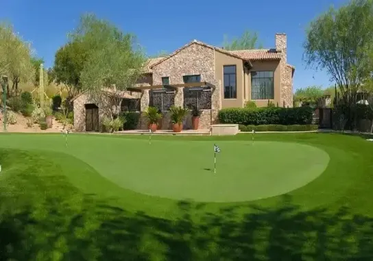A luxurious stone and stucco house with large glass windows is surrounded by trees. In the foreground, a pristine putting green reflects Avondale's Putting Green Experts' craftsmanship. Four flags flutter under the clear, blue sky that completes this serene setting.