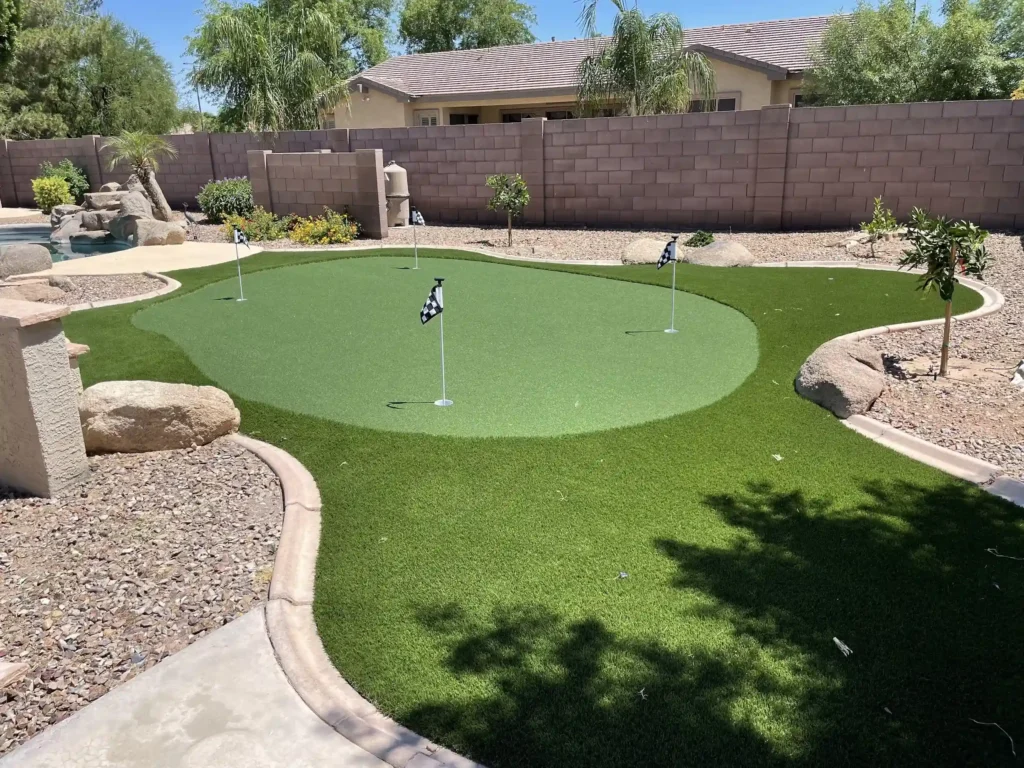 A backyard golf putting green designed by Chandler's Putting Green Experts features three small flags on artificial turf, surrounded by rocks and desert landscaping. A stone wall and a house enhance the beautiful setting under a clear blue sky.