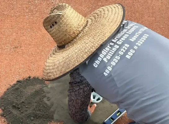 A person wearing a large straw hat and a gray shirt with Chandler's Putting Green Experts contact info on the back is kneeling, smoothing cement with a trowel. Nearby, a level tool rests on the reddish-brown surface, ensuring perfection in every detail.