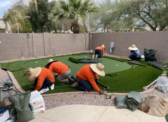 Four workers from Chandler's Putting Green Experts, wearing large hats and orange shirts, meticulously install artificial turf on a backyard putting green. They work on a gravel surface surrounded by trees and a stucco wall, with various tools and bags scattered around them.