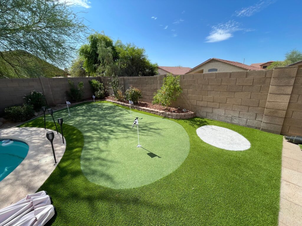 A backyard oasis crafted by Chandler's Putting Green Experts features a mini-golf green with a flagstick, encircled by low brick walls. A cozy round bunker area, lush landscaping with shrubs and trees, and a section of pool complete the scene under a clear blue sky.