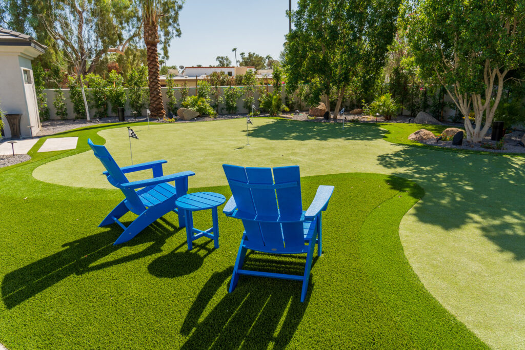 A backyard oasis with a mini putting green crafted by Chandler's Putting Green Experts features two blue Adirondack chairs and a matching table on the lush artificial grass. Trees and shrubs gracefully line the fenced perimeter, with a house partially visible on the left.
