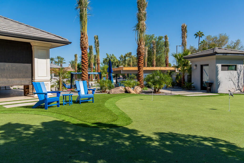 A backyard crafted by Chandler's Putting Green Experts features a mini golf area among palm trees and a small building. Two blue chairs and a table grace the grass, while a shaded patio and pool with lounge chairs bask under the clear blue sky.