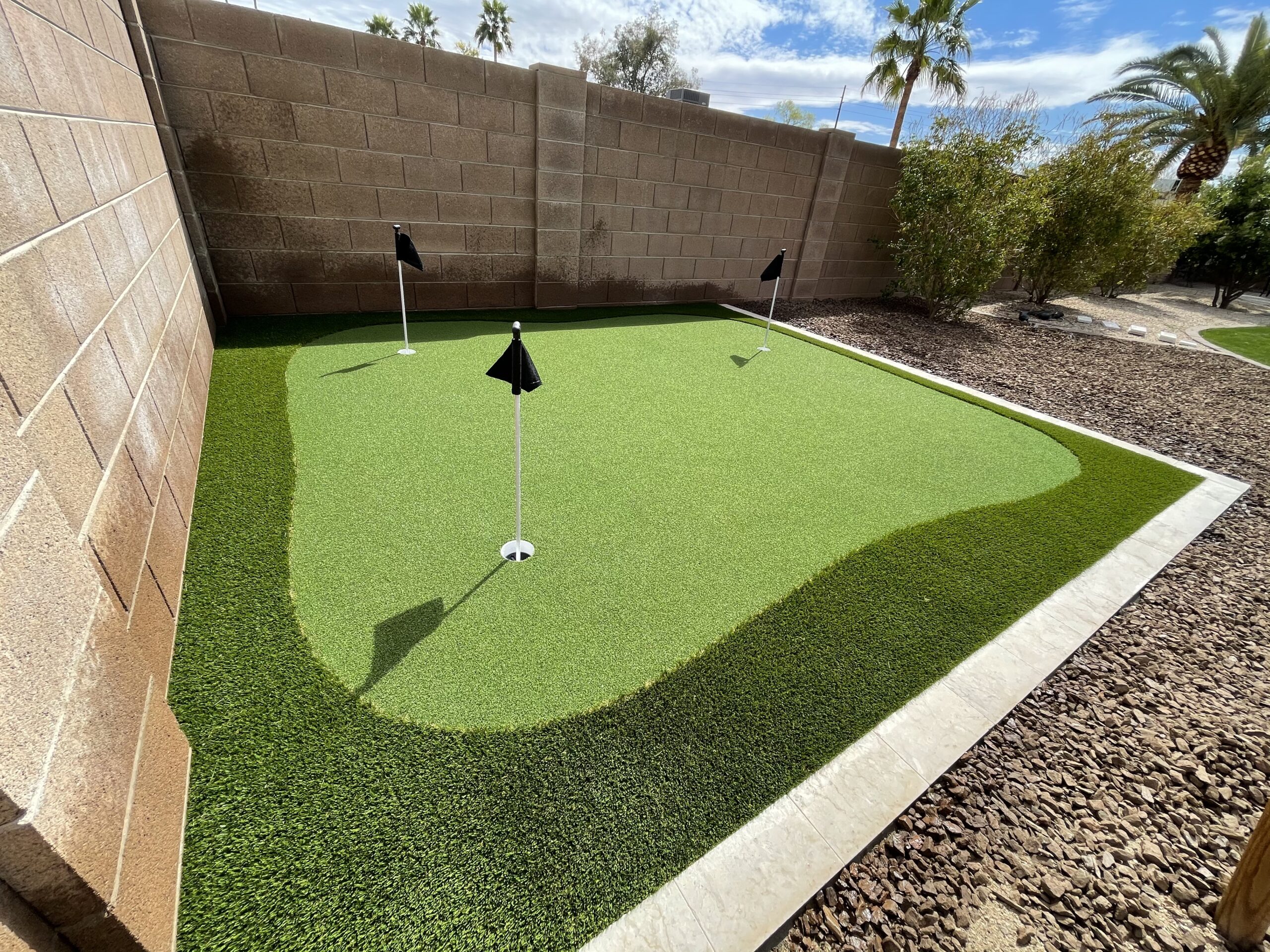 A small artificial putting green with three holes, crafted by Chandler's Putting Green Experts, is nestled in a backyard. It's framed by a brick wall and stone landscaping, with palm trees and bushes swaying gently under a partly cloudy sky.