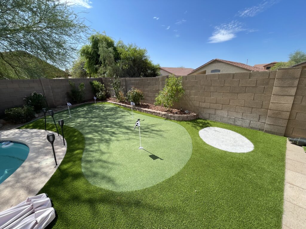 A backyard showcases a mini putting green crafted by Chandler's Putting Green Experts, featuring two white circles and small flags. It's encircled by a stone wall, trees, and shrubs. A glimpse of the pool and a sunny sky with scattered clouds complete the serene scene.