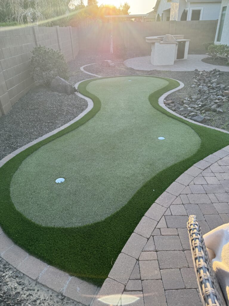 Transform your outdoor space with a backyard mini-golf putting green by Chandler's Putting Green Experts. Featuring two holes, surrounded by pebble landscaping and pavers, the yard is fenced with a stone wall and boasts a built-in grill area. The setting sun casts a warm glow over this serene haven.