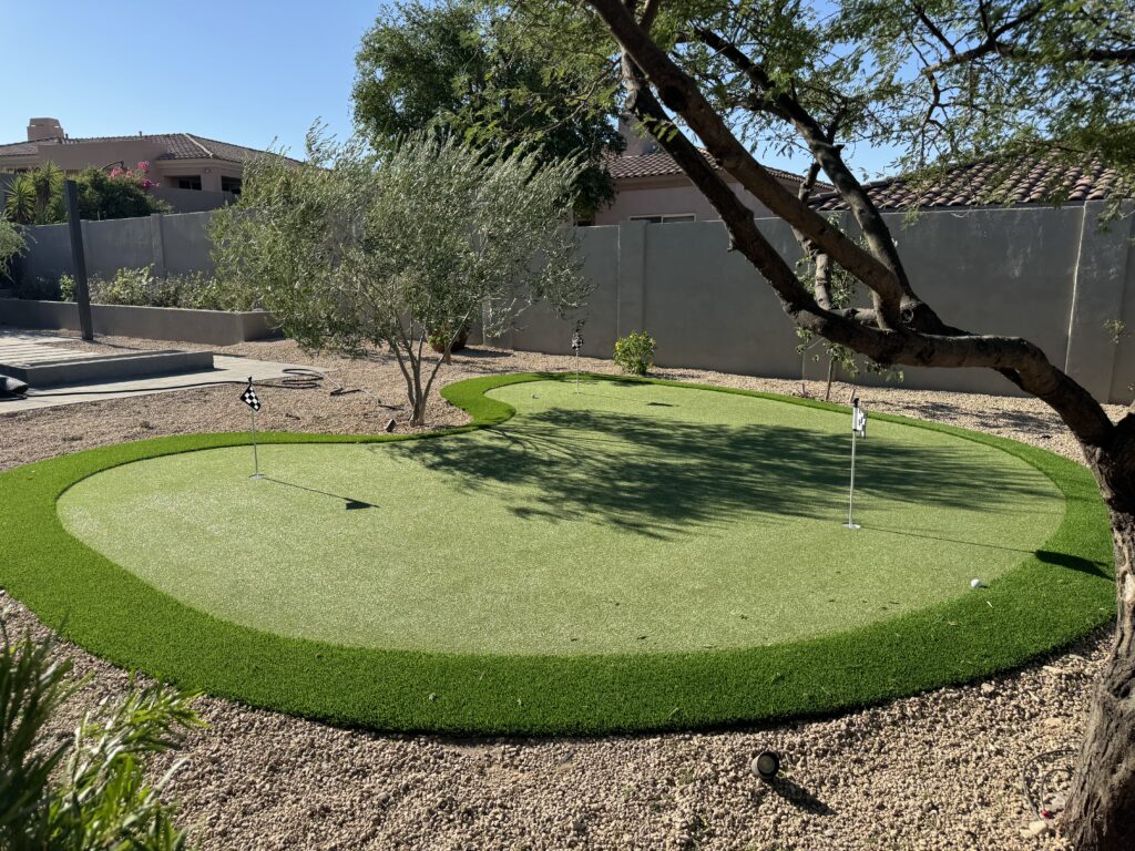 A backyard showcases a small putting green surrounded by gravel and sparse vegetation, crafted with the skill of Chandler's Putting Green Experts. Two small flags mark the neatly bordered green, enclosed by a beige wall with houses peeking through in the background.
