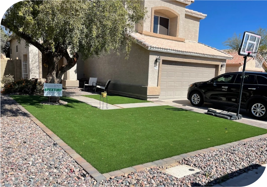 A suburban house with a two-car garage and beige exterior boasts a driveway lined with rocks. The front yard features a large tree and lush artificial grass, thanks to top-notch Artificial Grass Installation Services. A portable basketball hoop stands beside the black SUV parked there.