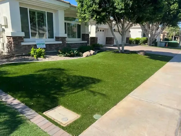 A suburban home with an Apex Turf manicured green lawn and a surrounding garden featuring bushes and rocks. The house has large windows with shutters, is partly clad in stone, and faces a sidewalk that runs in front.