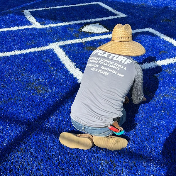 Apex Turf installer kneeling on blue artificial sports turf, wearing a straw hat and a gray shirt with Apex Turf logo on it. They diligently work on the sports field, bordered by crisp white lines.