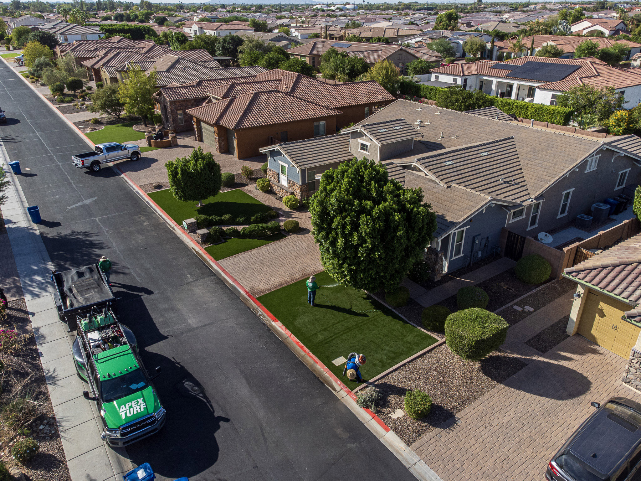 Aerial view of a suburban neighborhood with single-story homes, well-maintained lawns, and trees. Two people, possibly from Queen Creek's Artificial Grass Experts, are landscaping in a yard. A truck with equipment is parked on the street. The sky is clear and the setting is sunny.