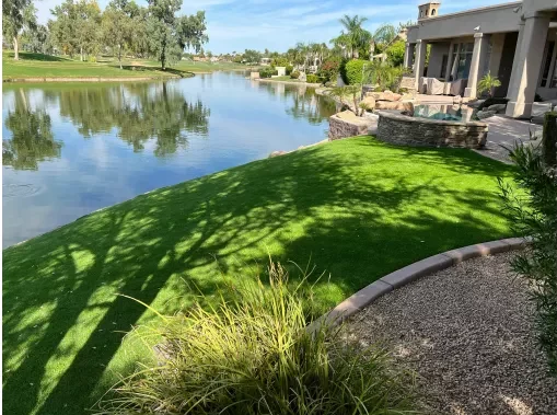 A serene landscape featuring a lush, green artificial grass Chandler lawn by Apex Turf beside a calm lake, bordered by trees and shrubs. A modern house with a patio overlooks the water. The sky is clear and blue, creating a peaceful atmosphere.