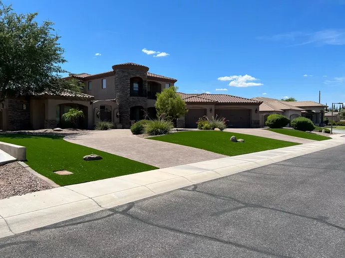 A large, modern house with a stone facade and tile roof sits on a suburban street. The front yard showcases Apex Turf's well-maintained artificial grass, complemented by small shrubs and a paved driveway. The sky is clear with a few scattered clouds over Chandler.